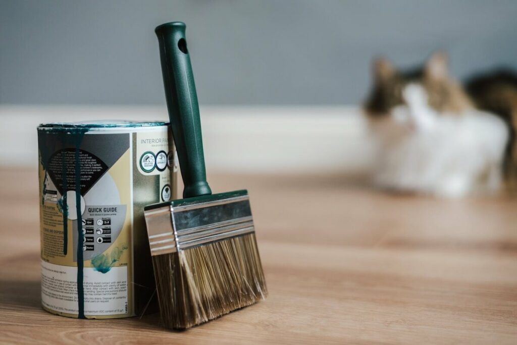 A can of paint and a brush on a wooden floor, with a cat behind